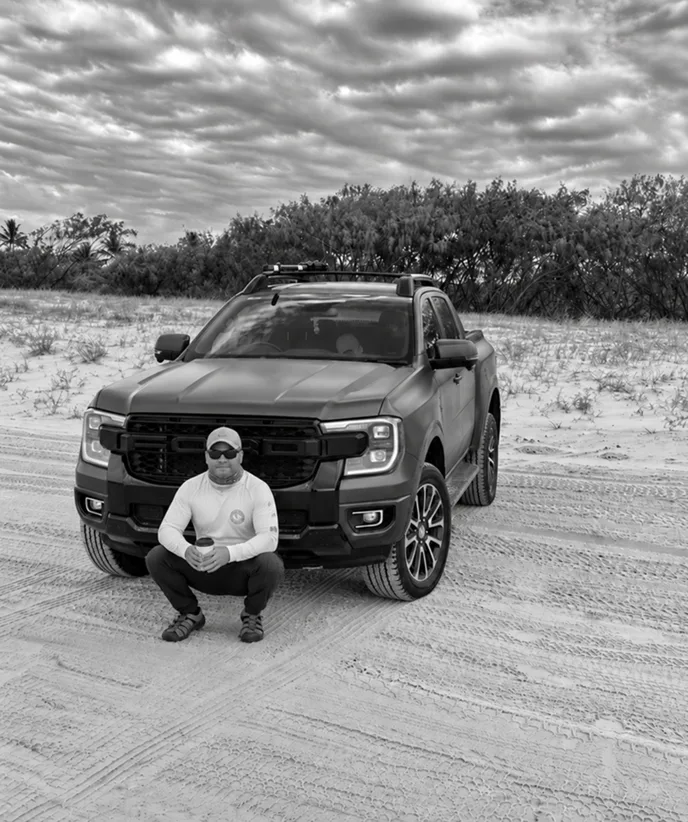 Greyscale photo of Tom crouching in front of his Ford Ranger on the sand dunes of K’gari
